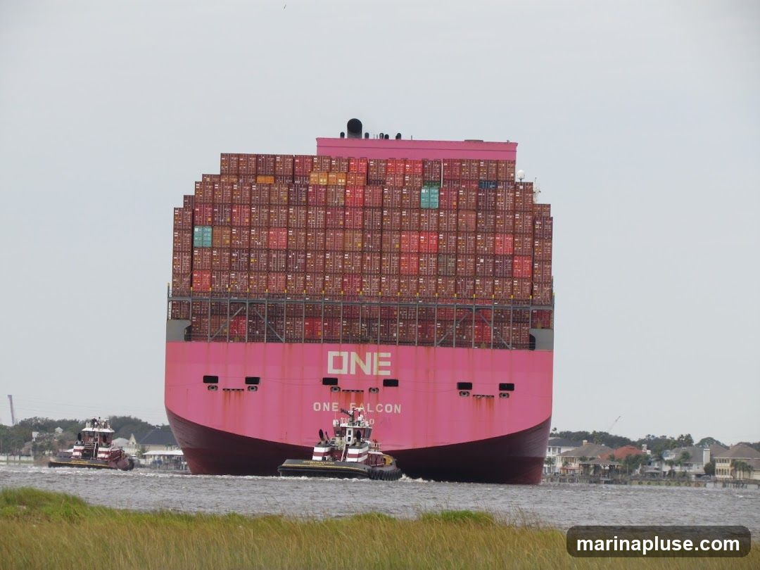 A large pink container ship, the ONE FALCON, is being guided by two tugboats through a body of water. The ship is laden with numerous stacked shipping containers, predominantly red, with some green and blue containers visible. The ship's name and logo are clearly visible on its stern. The background shows a shoreline with houses and trees under an overcast sky.