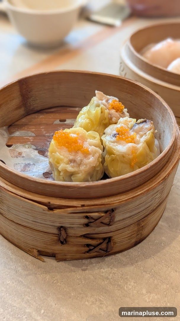 A close-up shot of three steamed dumplings in a bamboo steamer basket. The dumplings are topped with bright orange roe. In the background, out of focus, are more bamboo steamers and bowls.