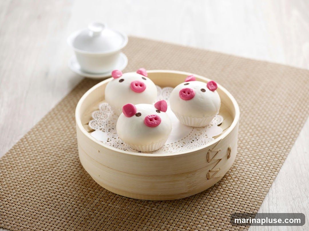 Three white steamed buns decorated to look like pig faces are presented in a bamboo steamer basket lined with a doily. Each bun has pink ears and snout, and small black eyes. A white teacup and saucer are visible in the background.