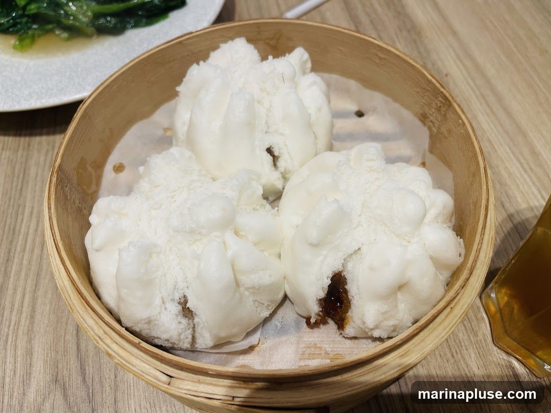 Three steamed buns are served in a bamboo steamer basket. One bun has a filling visible. In the background, a plate of green vegetables and a glass of amber liquid are present.