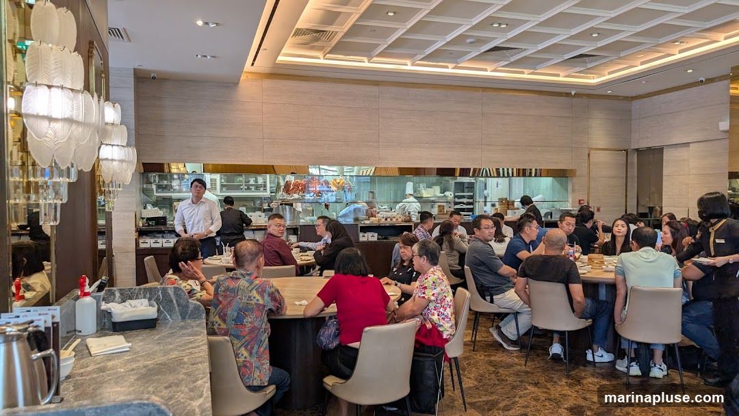 A busy restaurant interior with multiple tables occupied by diners. A partially visible open kitchen is in the background, showing chefs at work and hanging roasted meats. Elegant chandeliers hang from the ceiling.