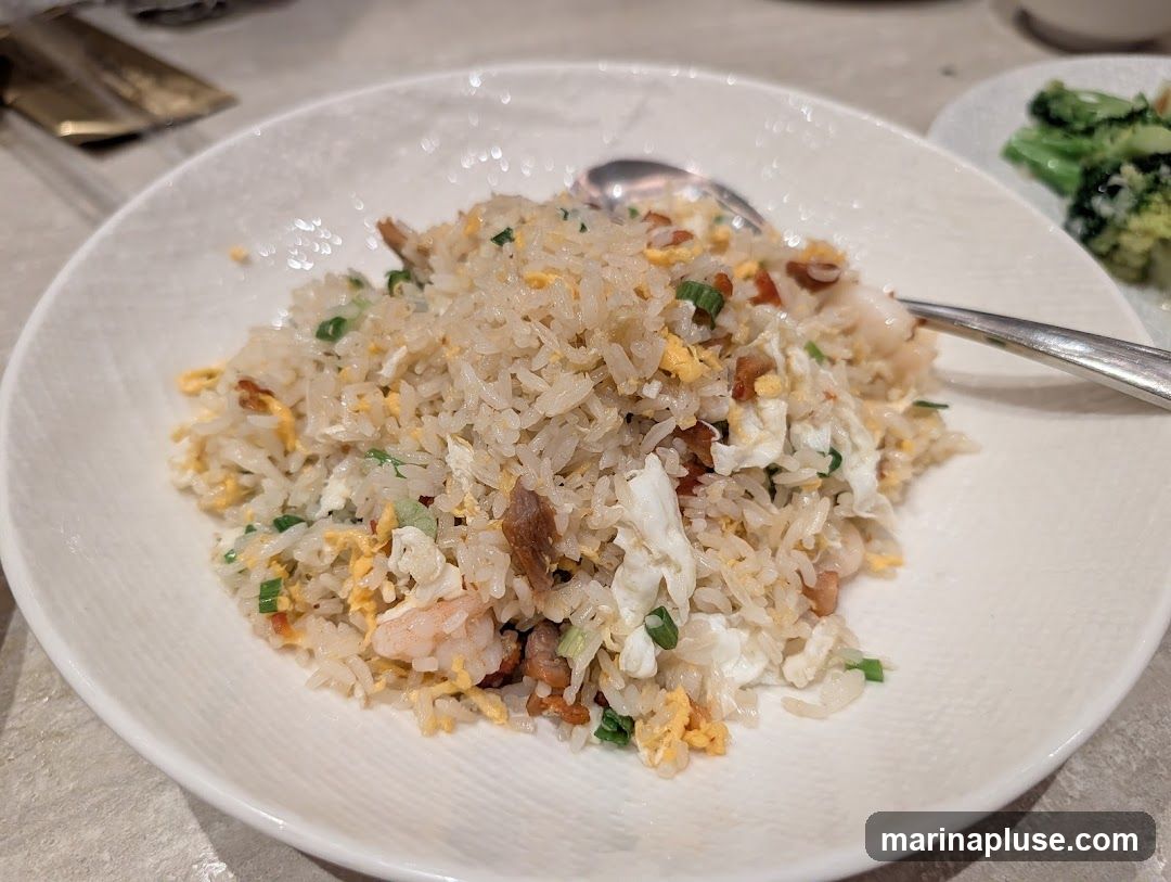 A close-up shot of a plate of shrimp fried rice, with visible pieces of shrimp, scrambled egg, and green onions mixed into the rice. A spoon rests on the side of the plate. In the background, a small plate of broccoli is visible.