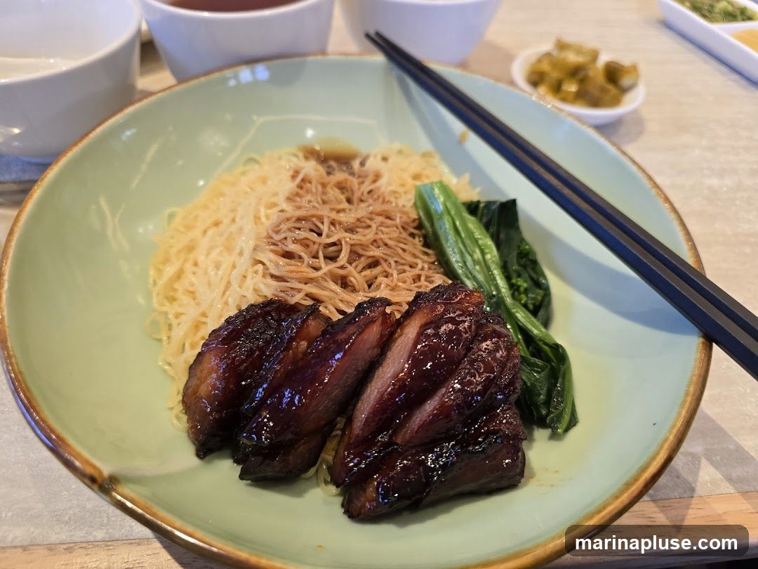 A close-up shot of a bowl of noodles topped with slices of glistening roast pork and a side of blanched greens. Chopsticks rest on the side of the bowl. In the background, there are two white bowls and a small dish of pickled vegetables.