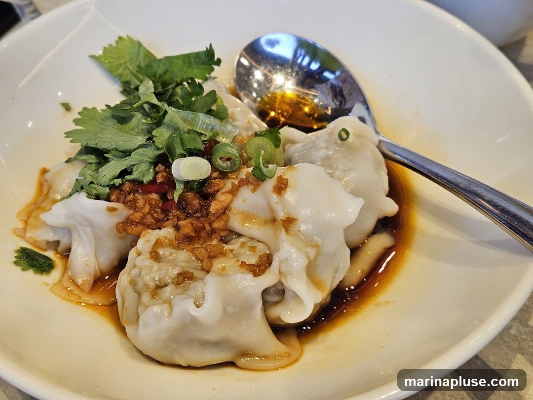 A close-up shot of a bowl of wontons in a spicy sauce, garnished with fresh cilantro and chopped scallions. A spoon rests in the bowl, reflecting the overhead lighting. The wontons are plump and appear to be filled with meat. The sauce is a rich, dark brown, with visible chili flakes and crispy garlic bits on top.