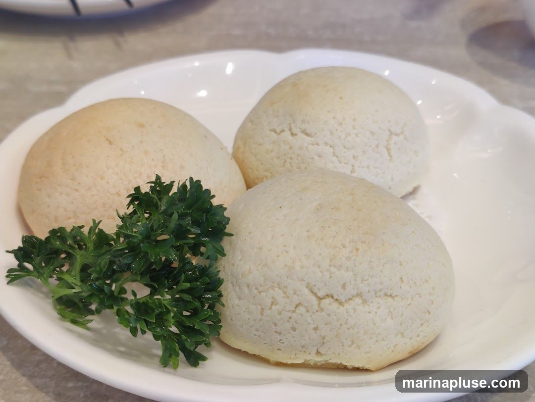 Three golden-brown baked buns are served on a white scalloped plate, garnished with a sprig of fresh parsley. The buns have a slightly cracked, textured surface and appear soft and fluffy.