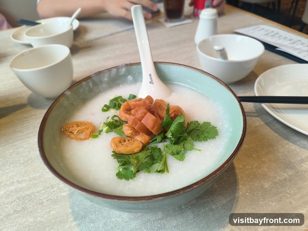 A close-up shot of a bowl of congee, a rice porridge, topped with chopped scallions, cilantro, and fried dough fritters. The bowl is light teal with a brown rim. There are several small white bowls and a spoon on the table, along with chopsticks and a menu in the background.