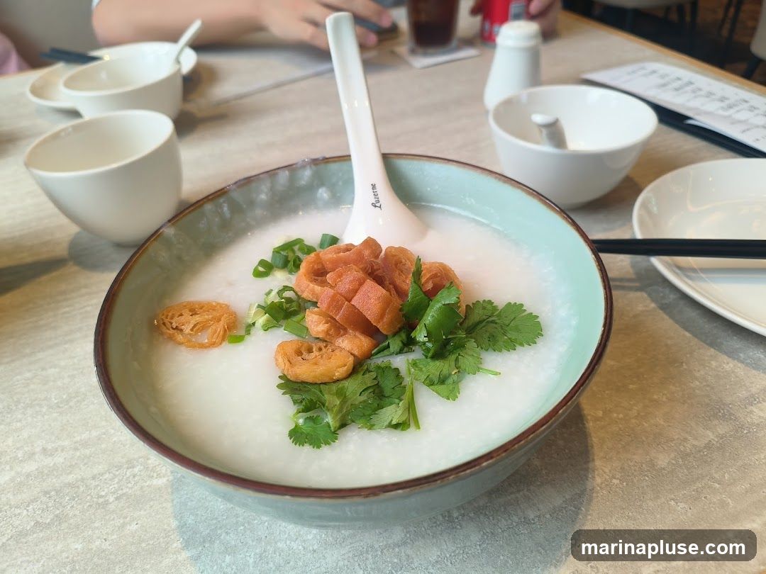 A close-up shot of a bowl of congee, a rice porridge, topped with chopped scallions, cilantro, and fried dough fritters. The bowl is light teal with a brown rim. There are several small white bowls and a spoon on the table, along with chopsticks and a menu in the background.