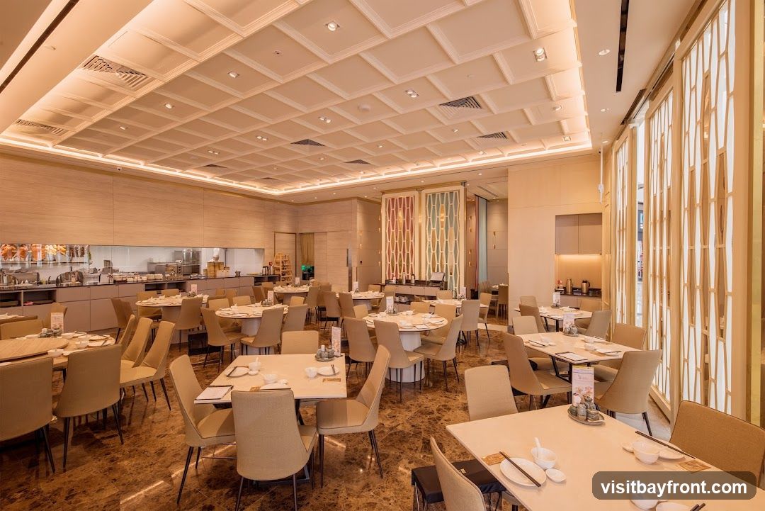 A spacious and elegantly decorated dining area in a restaurant. The room features numerous round and rectangular tables, each set with white tableware and chopsticks. The chairs are upholstered in a neutral beige fabric. The ceiling is intricately designed with a coffered pattern and recessed lighting. The walls are adorned with light wood paneling and decorative screens. In the background, a glimpse of the kitchen area is visible, with stainless steel appliances and a chef in uniform. The flooring is a polished marble with a rich brown hue.