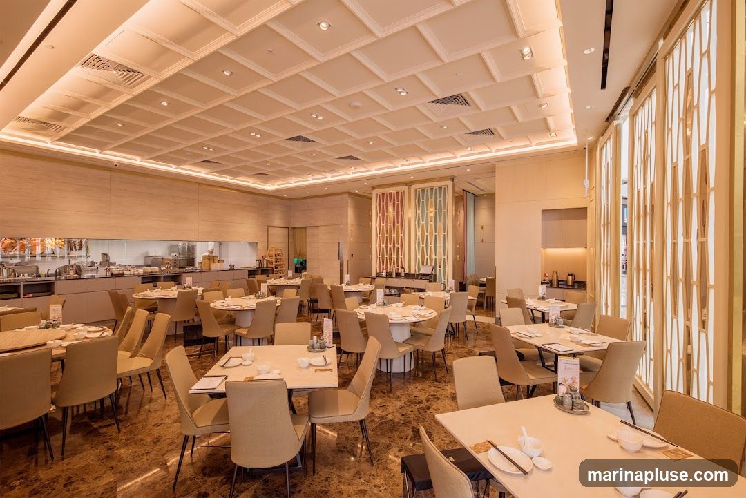 A spacious and elegantly decorated dining area in a restaurant. The room features numerous round and rectangular tables, each set with white tableware and chopsticks. The chairs are upholstered in a neutral beige fabric. The ceiling is intricately designed with a coffered pattern and recessed lighting. The walls are adorned with light wood paneling and decorative screens. In the background, a glimpse of the kitchen area is visible, with stainless steel appliances and a chef in uniform. The flooring is a polished marble with a rich brown hue.