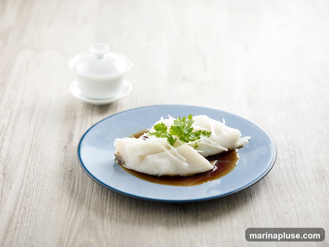 Two pieces of steamed fish fillets are served on a blue plate with a dark sauce and topped with shredded ginger and fresh cilantro. A small white lidded bowl sits in the background on a light wooden surface.