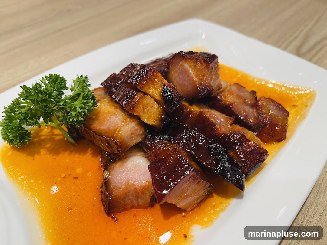 A plate of roasted pork belly, sliced and served with a glistening sauce and a sprig of parsley. The pork has a crispy, caramelized exterior and tender, juicy meat.
