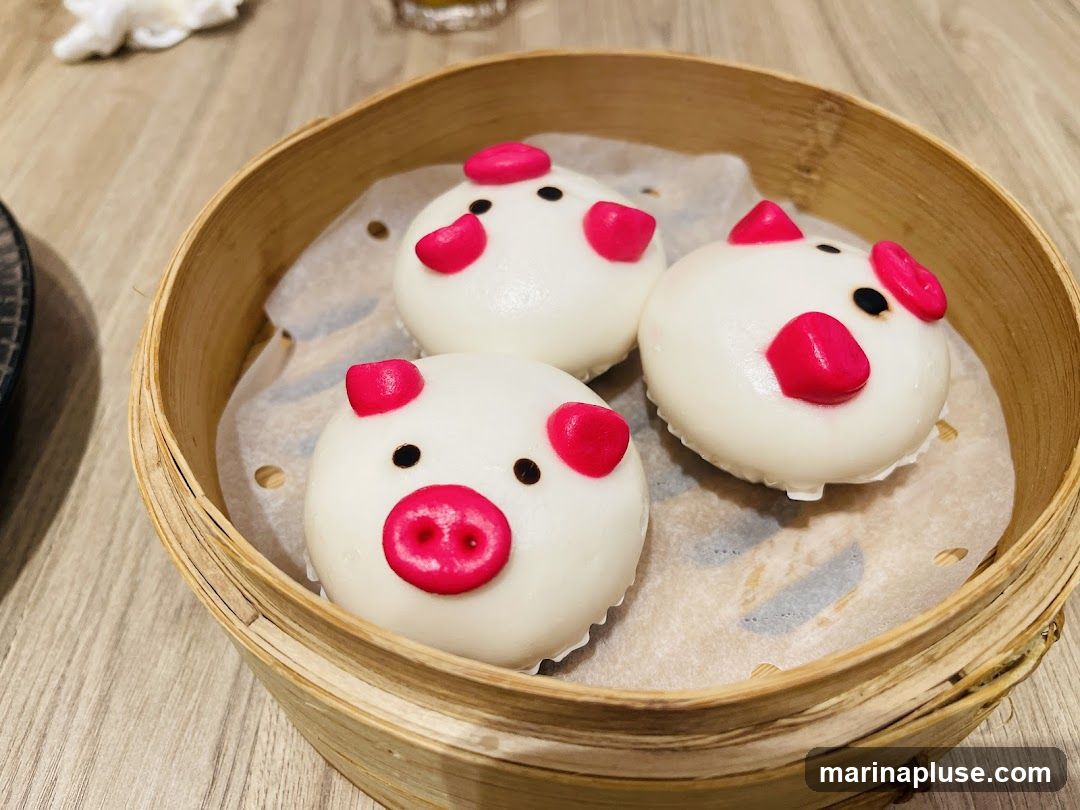 Three white steamed buns decorated to look like pig faces are presented in a bamboo steamer basket. Each bun has pink ears and snout, and black eyes.
