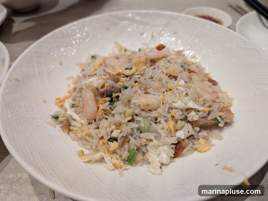 A close-up shot of a plate of shrimp fried rice. The rice is mixed with scrambled eggs, pieces of pork, and green onions. Several shrimp are visible throughout the dish. The plate has a textured rim.