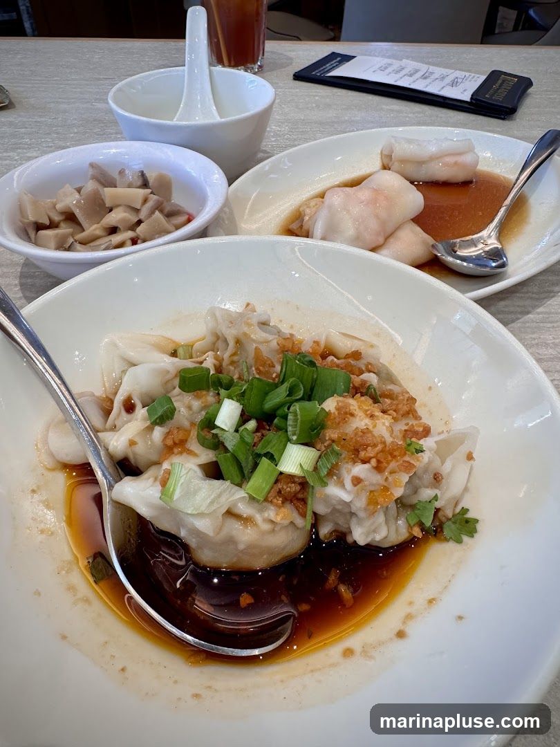 A close-up shot of a meal featuring several dishes. In the foreground, a white bowl holds a serving of dumplings in a dark sauce, garnished with chopped green onions and fried garlic bits. A spoon rests in the bowl. To the left, a smaller white bowl contains a pickled appetizer. In the background, another white oval dish holds several pieces of shrimp-filled rice noodle rolls in a light brown sauce, with a spoon. A glass of iced tea and a menu are visible on the table.