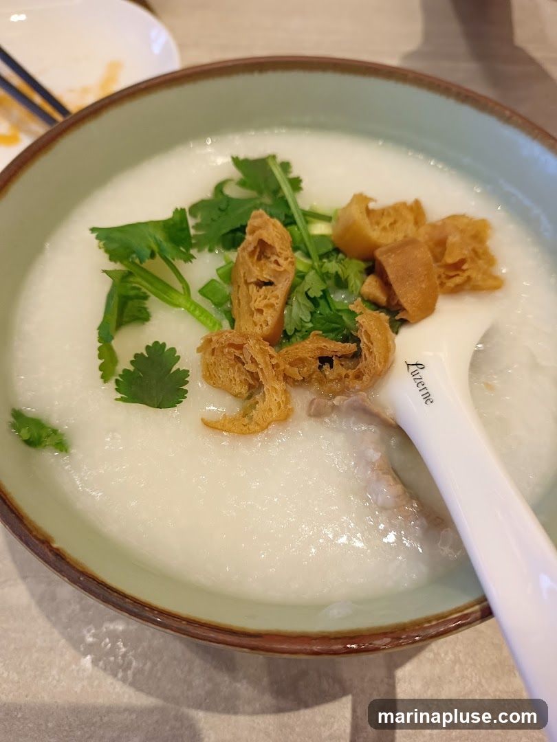 A close-up shot of a bowl of congee, a rice porridge, topped with chopped cilantro, green onions, and fried dough fritters. A piece of meat is visible beneath the surface of the congee. A white spoon with the word "Luzerne" printed on it rests in the bowl. In the background, a partially eaten dish and chopsticks are visible.