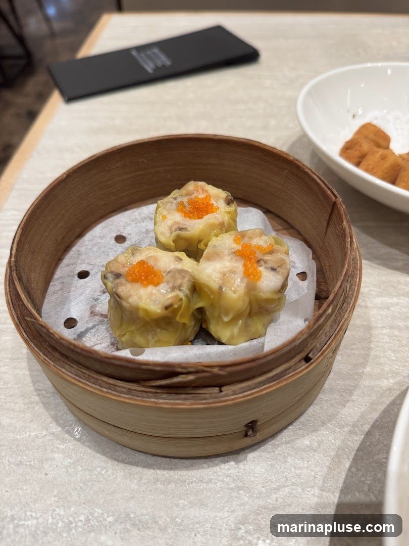 A close-up shot of three steamed dumplings, likely siu mai, served in a bamboo steamer basket lined with white paper. The dumplings are topped with bright orange roe. In the background, a white bowl contains fried cubes, possibly tofu or bread, and a black menu or placemat is visible on the table.