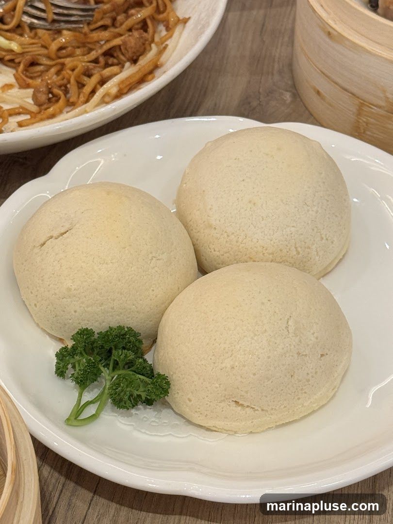 Three golden-brown steamed buns are presented on a white plate, garnished with a sprig of parsley. In the background, a plate of noodles with meat and vegetables is visible, along with a bamboo steamer.