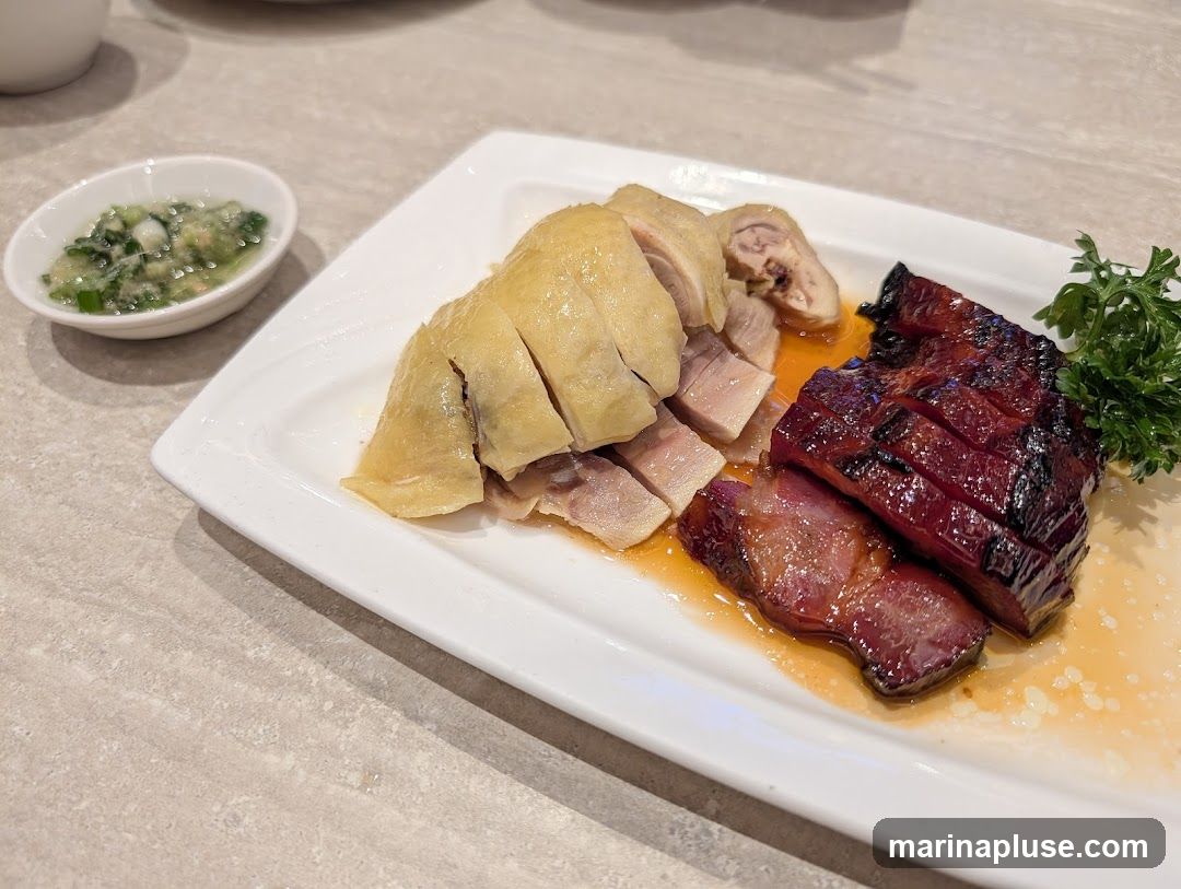 A white platter holds sliced roasted chicken and char siu (barbecued pork). A small dish of dipping sauce with chopped green onions and ginger is to the left. A sprig of parsley garnishes the char siu. The food is served on a textured, light-colored surface.