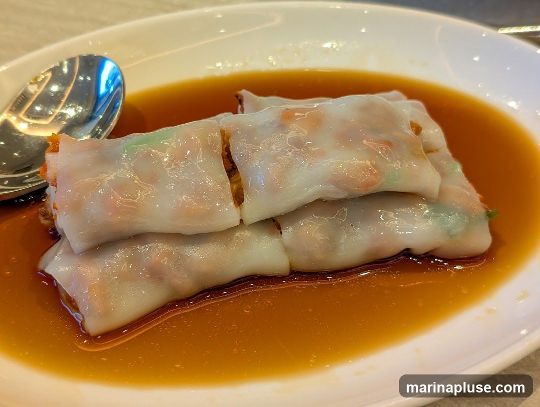 A close-up shot of shrimp rice noodle rolls served in a savory sauce on a white oval plate. A spoon is visible on the left side of the plate. The translucent rice noodle wrappers reveal the pink shrimp and green chives inside. The dish is glistening with the amber-colored sauce.