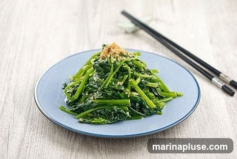 A plate of stir-fried green vegetables, possibly water spinach or similar, is presented on a light blue plate. The vegetables are piled high and topped with a sprinkle of toasted garlic or breadcrumbs. A pair of black chopsticks rests on a chopstick rest in the background, positioned to the right of the plate. The dish is set against a light-colored, textured surface, likely a wooden table.
