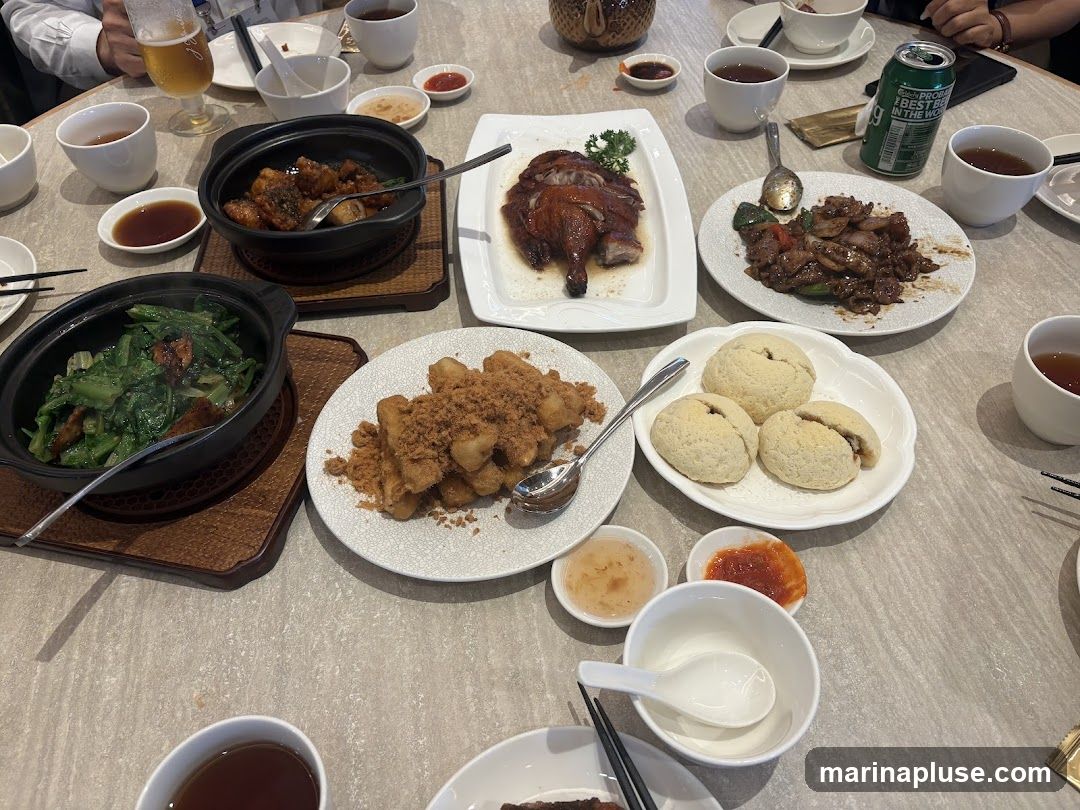 A table laden with various Chinese dishes, including roasted duck, stir-fried vegetables, meat dishes, and fried items. Several cups of tea and small dipping sauce bowls are also visible, along with a can of beer. The meal appears to be a shared dining experience.