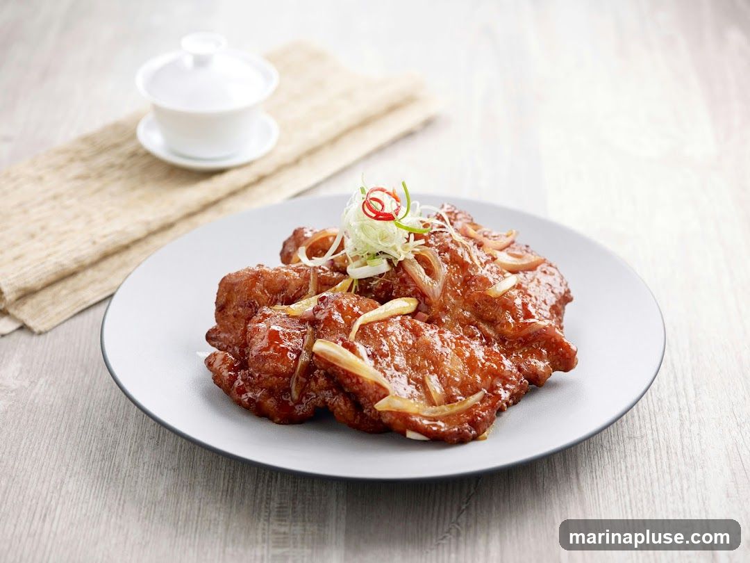 A plate of sweet and sour pork, garnished with thinly sliced scallions and chili peppers, is presented on a light gray plate. A small white tea cup and saucer sit on a beige cloth in the background. The dish is arranged on a light-colored wooden surface.