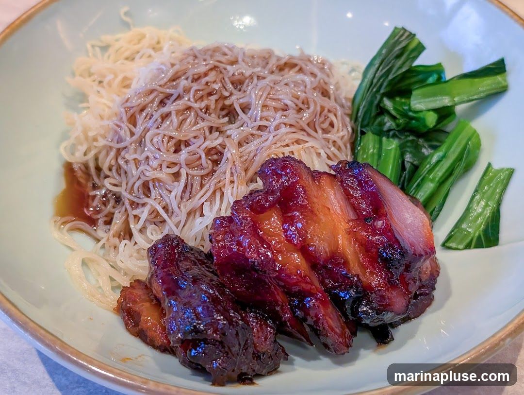 A close-up shot of a bowl of noodles topped with glistening char siu (barbecued pork) and a side of blanched green vegetables. The noodles are thin and appear to be tossed in a savory sauce, while the char siu has a deep reddish-brown glaze and visible layers of fat and meat. The greens are bright and fresh.