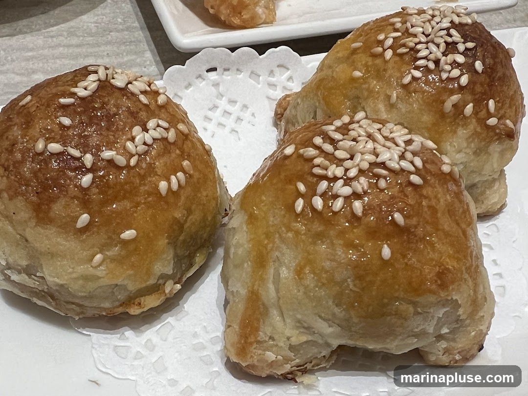 Three golden-brown pastries topped with sesame seeds are arranged on a white doily. The pastries appear to be baked and have a flaky texture.