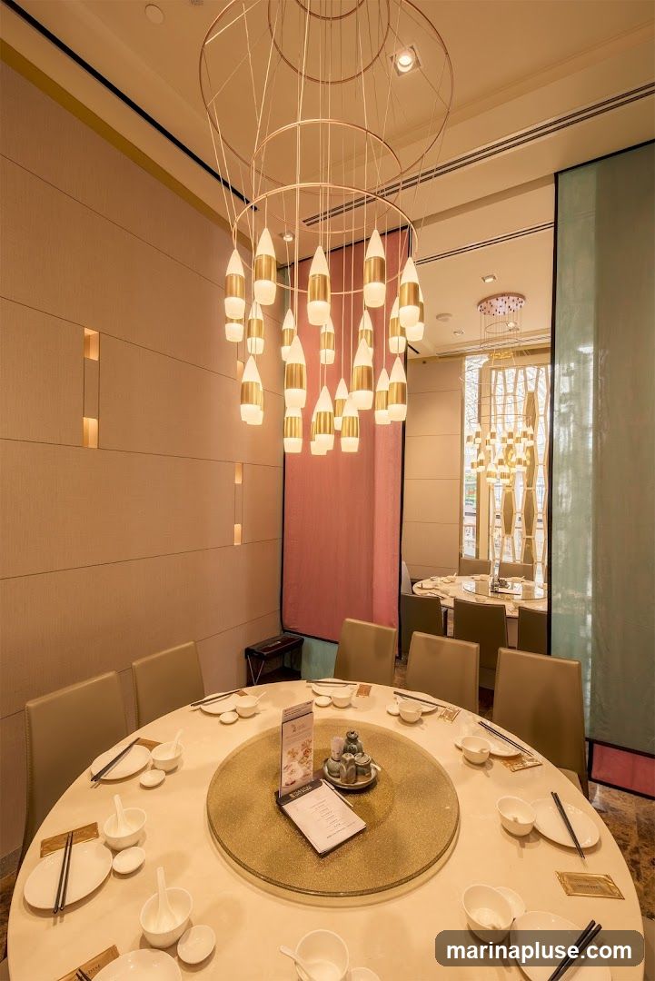 A well-lit dining room with multiple round tables set for meals. A large, modern chandelier with gold and white accents hangs from the ceiling, illuminating the space. The walls are textured in a neutral tone, with subtle vertical lighting elements. Decorative fabric panels in pink and teal add a touch of color. The tables are covered with white tablecloths and set with white plates, bowls, chopsticks, and other tableware. A menu is visible on one of the tables. In the background, other dining tables and chandeliers can be seen, suggesting a spacious restaurant interior.