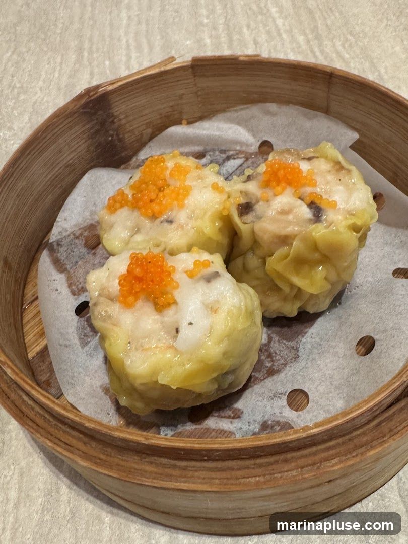 Three pieces of siu mai, a type of dim sum, are presented in a traditional bamboo steamer lined with parchment paper. Each siu mai is topped with bright orange roe. The steamer is placed on a textured surface, possibly a table.