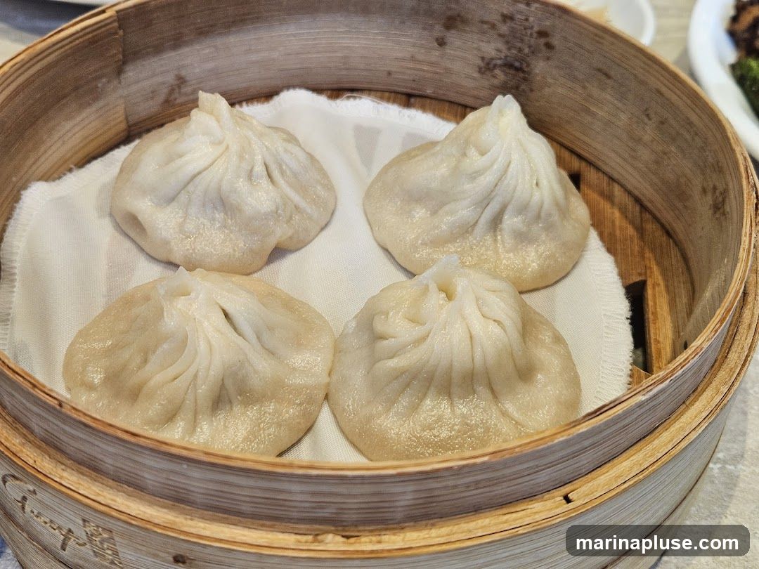 Four soup dumplings are presented in a bamboo steamer lined with white cloth. The dumplings are plump and have delicate pleats at the top.