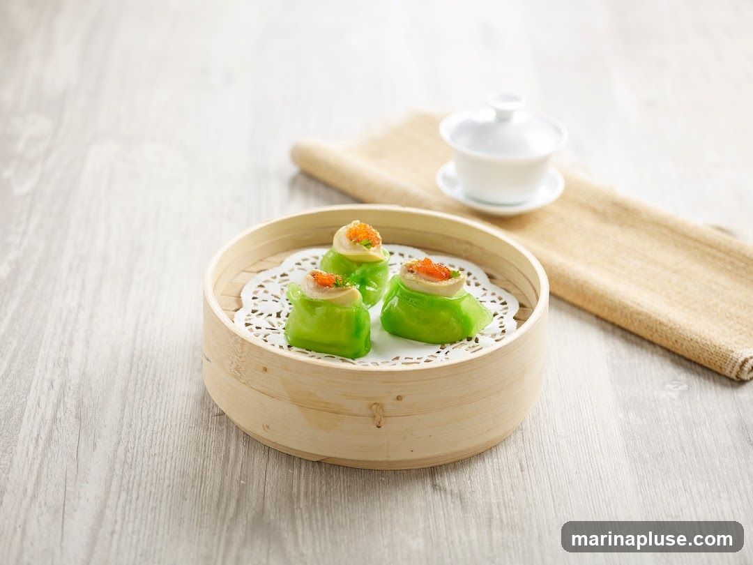 Three vibrant green dumplings are presented in a bamboo steamer lined with a white doily. Each dumpling is topped with a small dollop of filling and bright orange roe. A white tea cup and saucer sit on a folded beige cloth in the background, all arranged on a light-colored wooden surface.