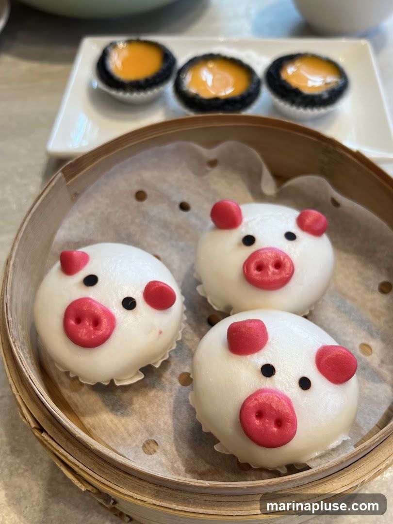 Three steamed buns shaped like pig faces are presented in a bamboo steamer. Behind them, on a white rectangular plate, are three small egg tarts with a dark crust and a bright orange filling.