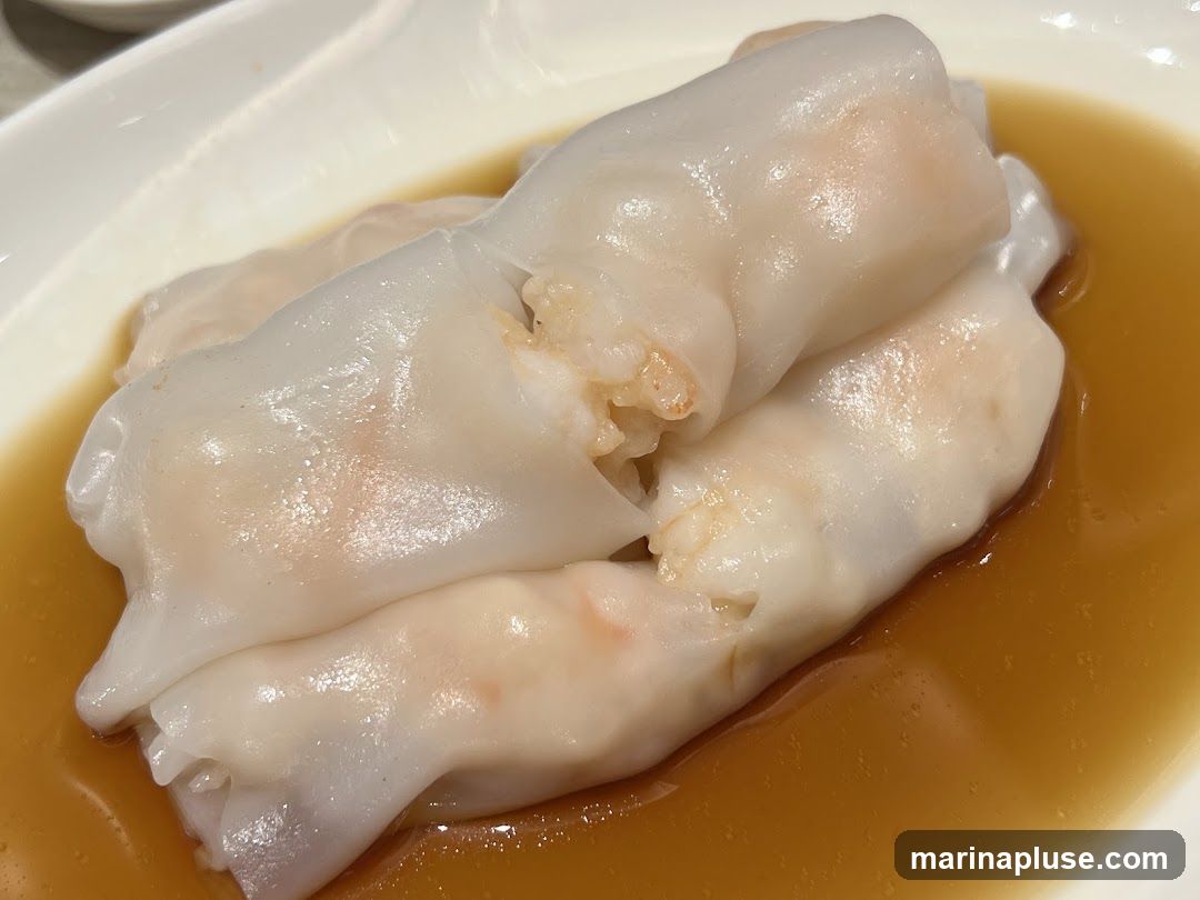 A close-up shot of three shrimp rice noodle rolls served in a white dish with a light brown sauce. The translucent rice noodle wrappers reveal pieces of pink shrimp inside. The dish appears to be a dim sum item.