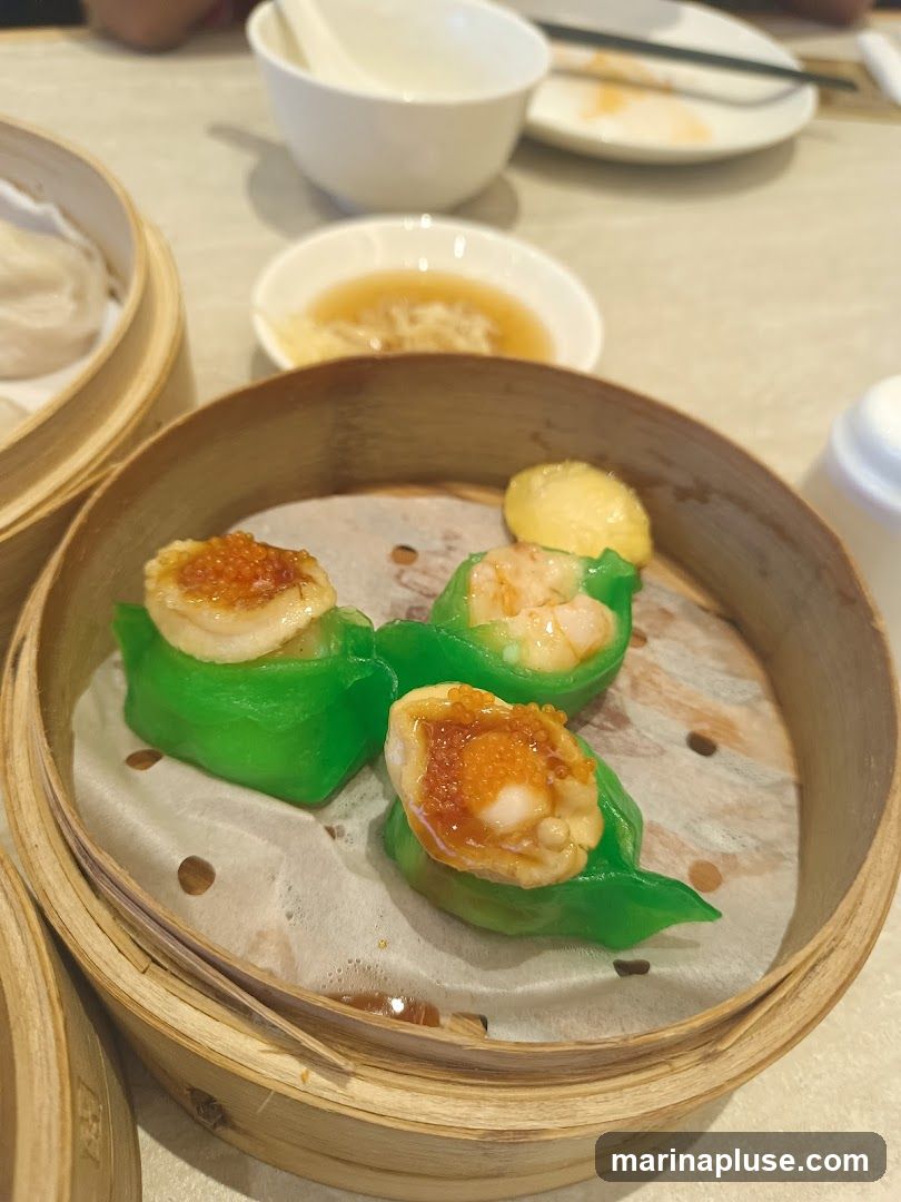 A close-up shot of a bamboo steamer basket filled with vibrant green dim sum dumplings. Each dumpling is topped with a piece of scallop and bright orange roe, and some contain shrimp. In the background, another steamer basket with white dumplings and a small dish of dipping sauce are visible on a table setting.