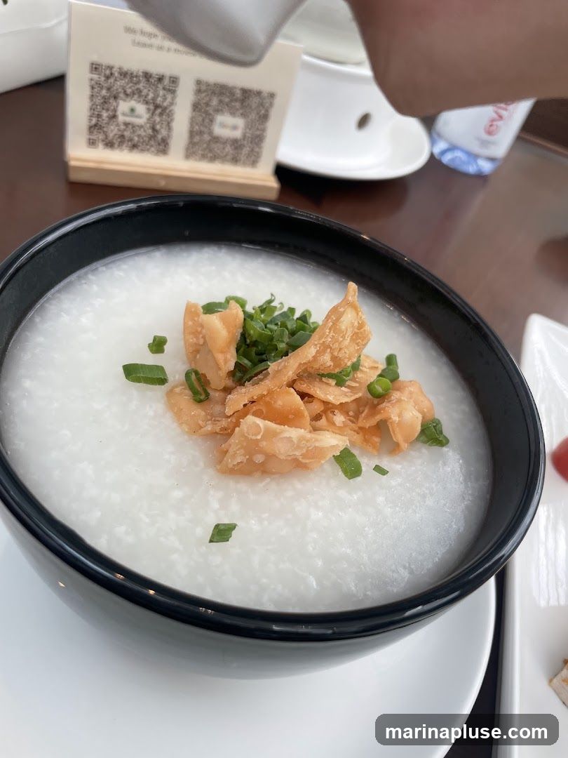 A close-up shot of a bowl of congee, a type of rice porridge, topped with chopped green onions and crispy fried wonton strips. The bowl is black with a white rim and sits on a white saucer. In the background, a small wooden stand holds two QR codes. A bottle of Evian water is also visible.