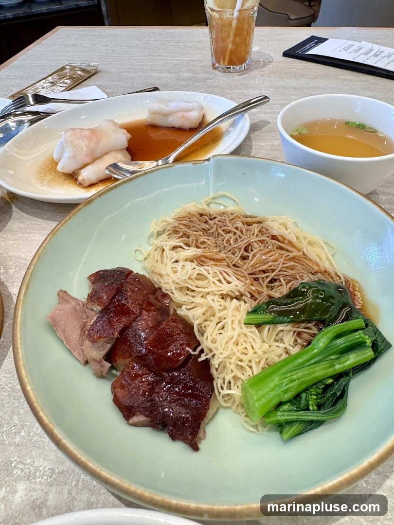 A plate of roast duck with noodles and greens is served with a side of soup and rice noodle rolls. A glass of iced tea is also on the table.