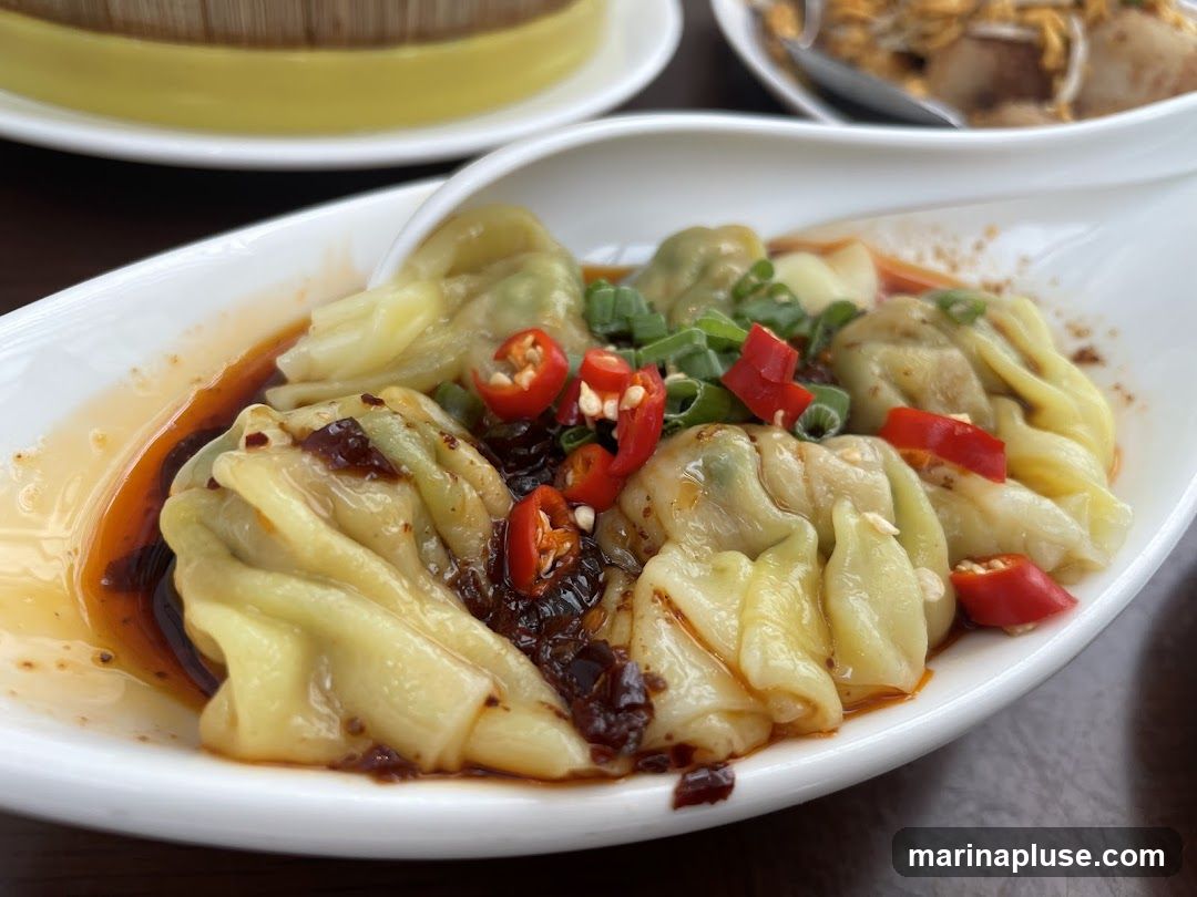 A close-up shot of wontons in a spicy chili oil sauce, garnished with chopped green onions and sliced red chilies. The wontons are light yellow and appear to be filled. In the background, out of focus, are other dishes, including what looks like a dim sum steamer basket and another plate of food.