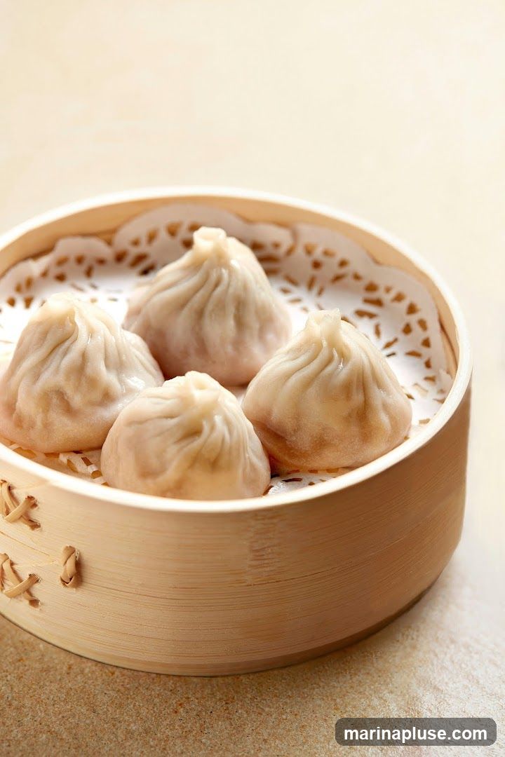 A close-up shot of four steamed dumplings in a bamboo steamer lined with a doily. The dumplings are plump and have delicate pleats at the top. The steamer is made of light-colored wood with woven accents. The background is a soft, neutral color.