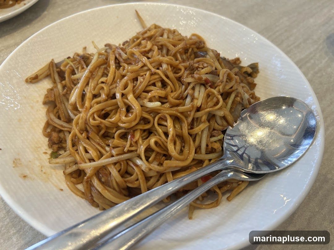 A close-up shot of a plate of noodles, likely a stir-fried dish, served with two spoons. The noodles are thin and coated in a savory sauce, mixed with bean sprouts and small pieces of meat or vegetables. The plate has a textured white rim.