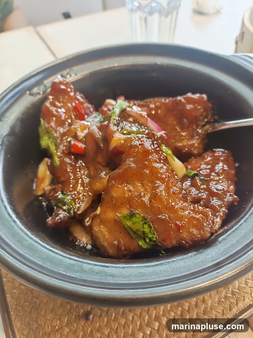 A close-up shot of a braised dish in a dark bowl. The dish appears to be meat, possibly fish or chicken, coated in a glossy, dark sauce with visible pieces of red chili, green herbs, and red onion. The food is served in a dark, textured bowl, placed on a woven placemat. A glass of water is visible in the background.