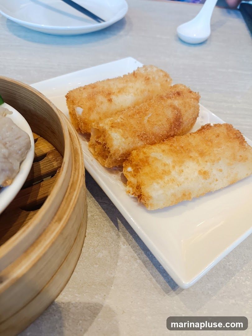 Three golden-brown fried spring rolls are served on a white rectangular plate. To the left, a bamboo steamer basket contains what appears to be dim sum. The table surface is a light-colored, textured material.