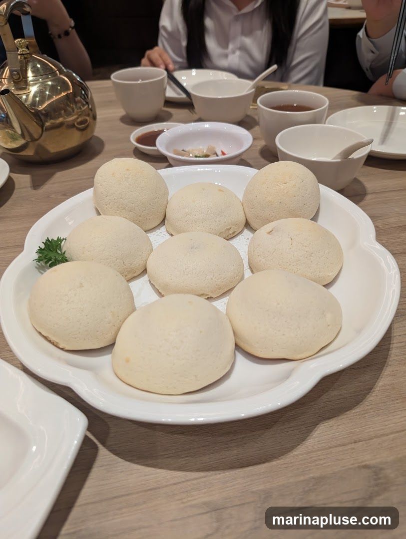 A white plate holds nine golden-brown baked buns, arranged in three rows. A small sprig of green parsley garnishes the left side of the plate. In the background, a golden teapot sits on a wooden table, alongside white teacups, small bowls of dipping sauce and pickled vegetables, and other tableware. Several people are seated at the table, partially visible.