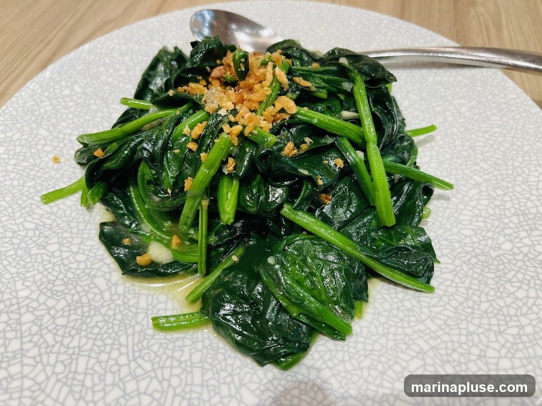 A close-up shot of sautéed spinach with garlic and crispy fried garlic bits on top, served on a textured white plate with a spoon in the background.