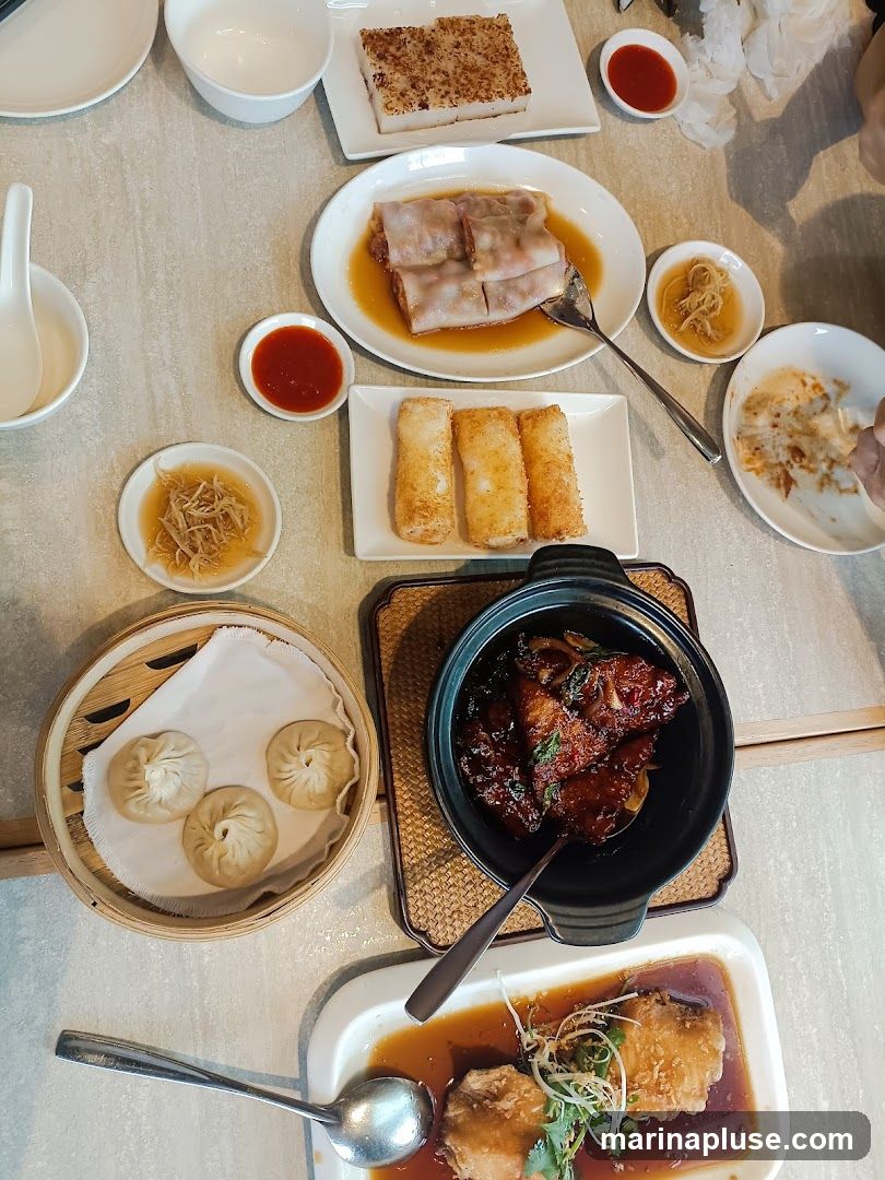 A table laden with various dim sum dishes, including steamed dumplings, rice noodle rolls, fried spring rolls, turnip cake, and braised meat. Small bowls of dipping sauce and pickled ginger are also visible.