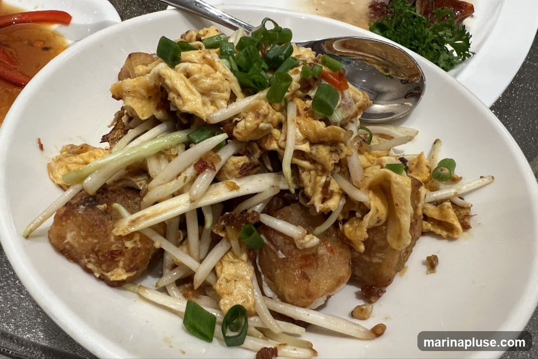 A close-up shot of a fried oyster omelet served in a white bowl. The dish is garnished with chopped green onions and appears to be made with oysters, scrambled eggs, and bean sprouts. Other dishes are visible in the background.