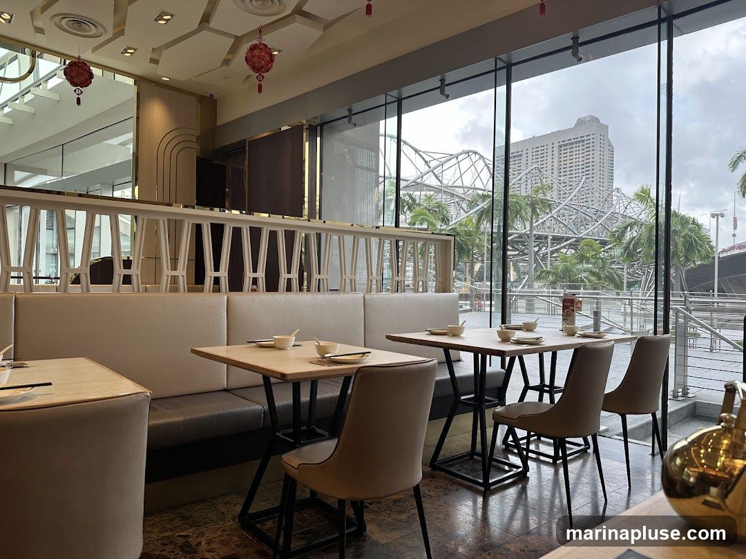 The image shows the interior of a restaurant with a view of a city skyline through large windows. The dining area features booth seating and tables set with bowls and chopsticks. Decorative lanterns hang from the ceiling. Outside, palm trees and a complex metal structure are visible, along with a large building in the distance.