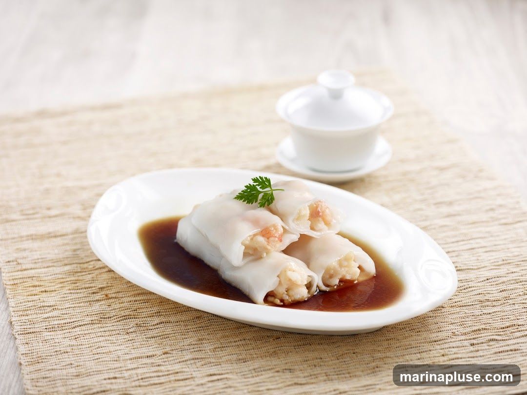 A close-up shot of three shrimp rice noodle rolls served in a dark sauce on a white oval plate. The rolls are translucent, revealing the pink shrimp filling. A sprig of parsley garnishes the top roll. A small white lidded bowl sits in the background on a woven placemat.