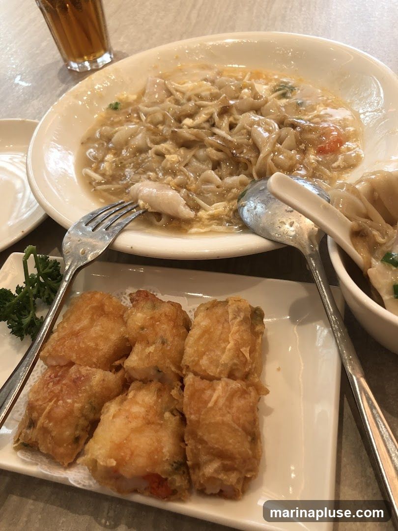 A close-up shot of a meal featuring fried rolls and a noodle dish. In the foreground, a white rectangular plate holds six golden-brown fried rolls, some with visible shrimp filling. A fork rests on the plate. Behind the rolls, a large white oval bowl contains a noodle dish with a creamy sauce, pieces of meat, and some vegetables. A spoon and a white utensil are in the bowl. To the left, a glass of amber liquid, likely tea, is partially visible. In the background, a white bowl contains dumplings.