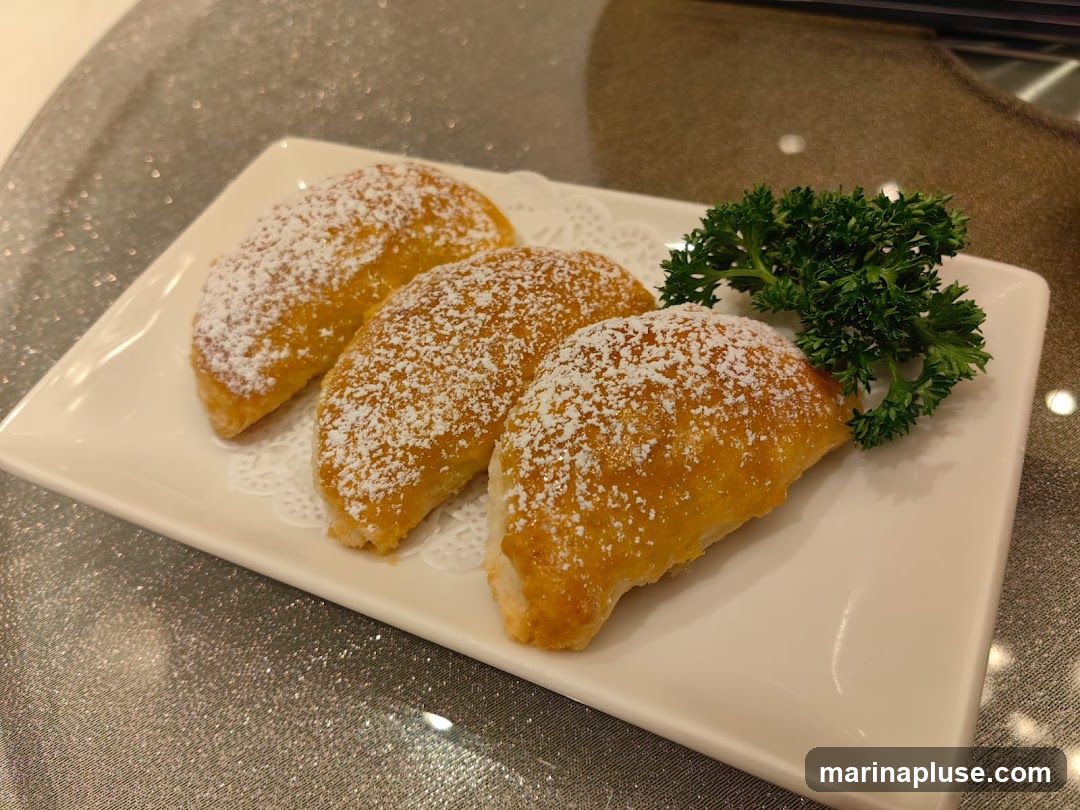 Three golden-brown pastries, dusted with powdered sugar, are arranged on a white rectangular plate. A sprig of fresh parsley garnishes the side of the plate. The pastries appear to be crescent-shaped and are served on a table with a textured, shimmering tablecloth.